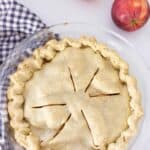 homemade apple pie in a glass pie dish on top antique stove with a blue and white checked towel under the pie and two apples in the back