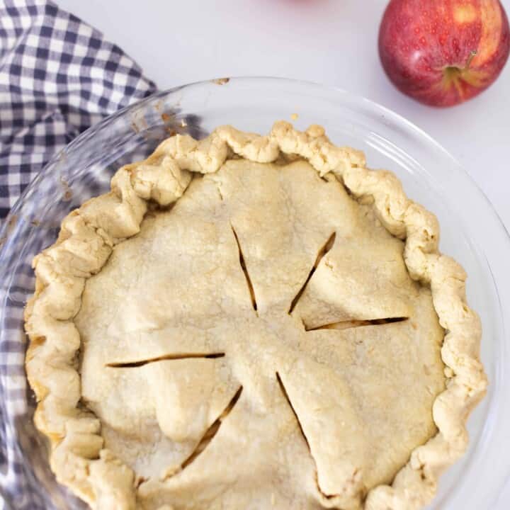 homemade apple pie in a glass pie dish on top antique stove with a blue and white checked towel under the pie and two apples in the back