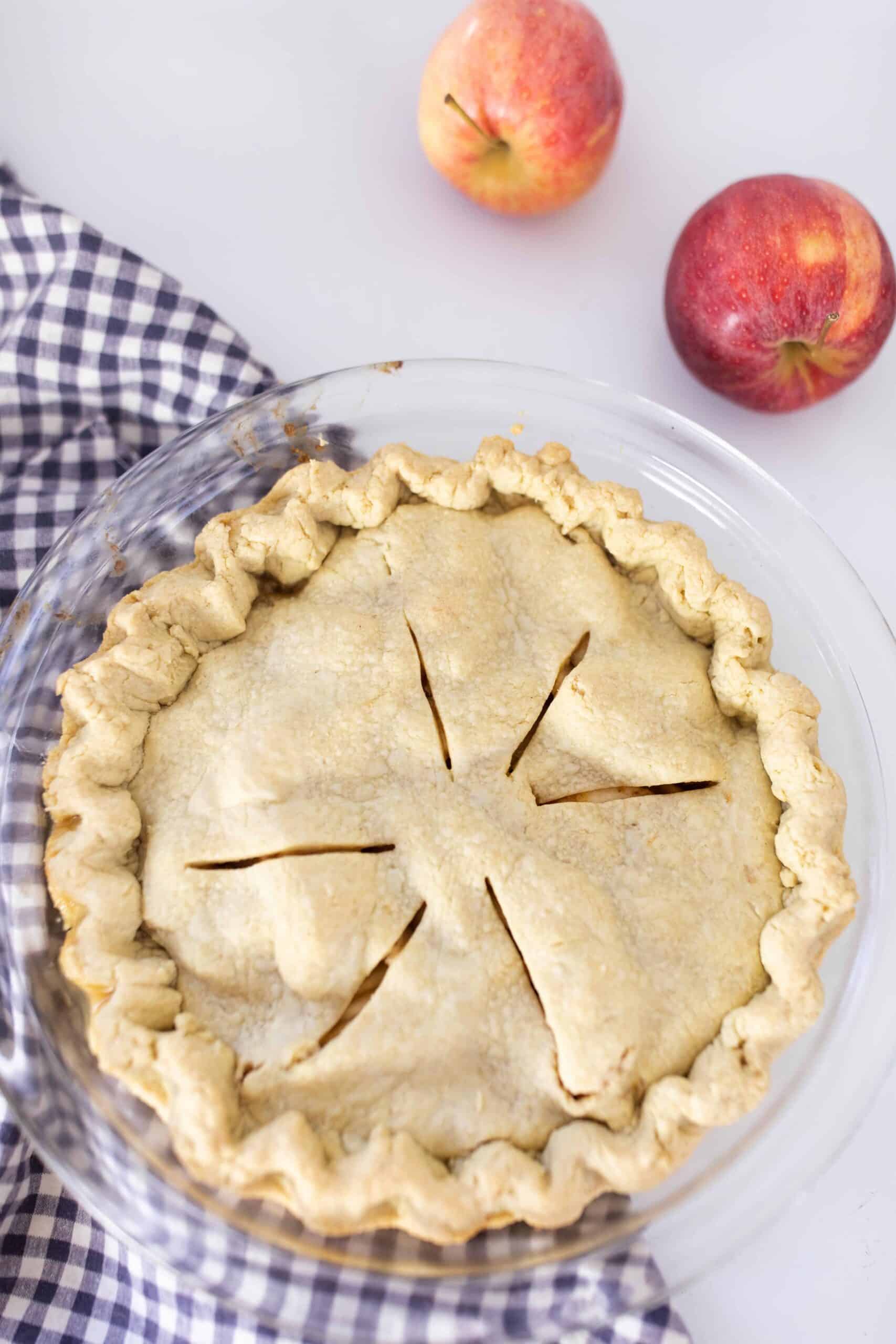 homemade apple pie in a glass pie dish on top antique stove with a blue and white checked towel under the pie and two apples in the back