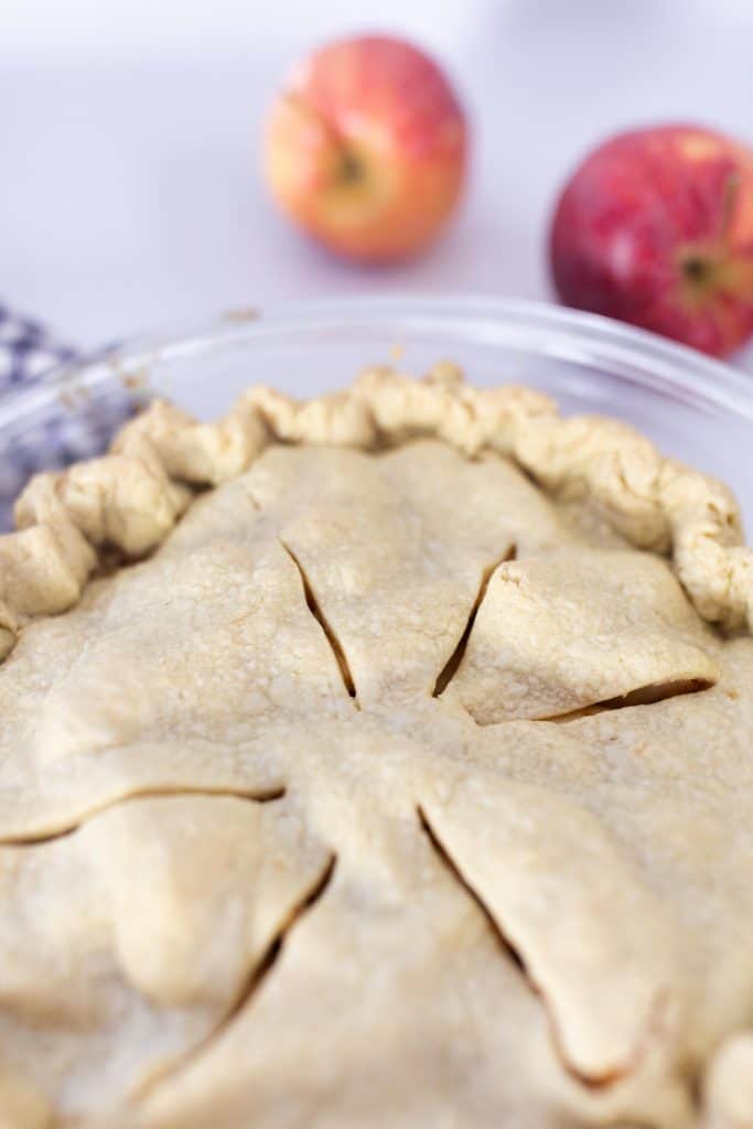 close up picture of the top of a homemade apple pie in a glass pie plate with apples in the background