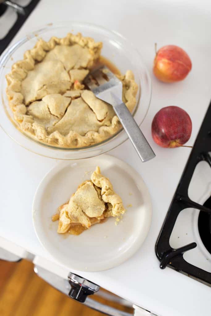 apple pie on a vintage stove with a slice on a white plate. Apples to left of the pie