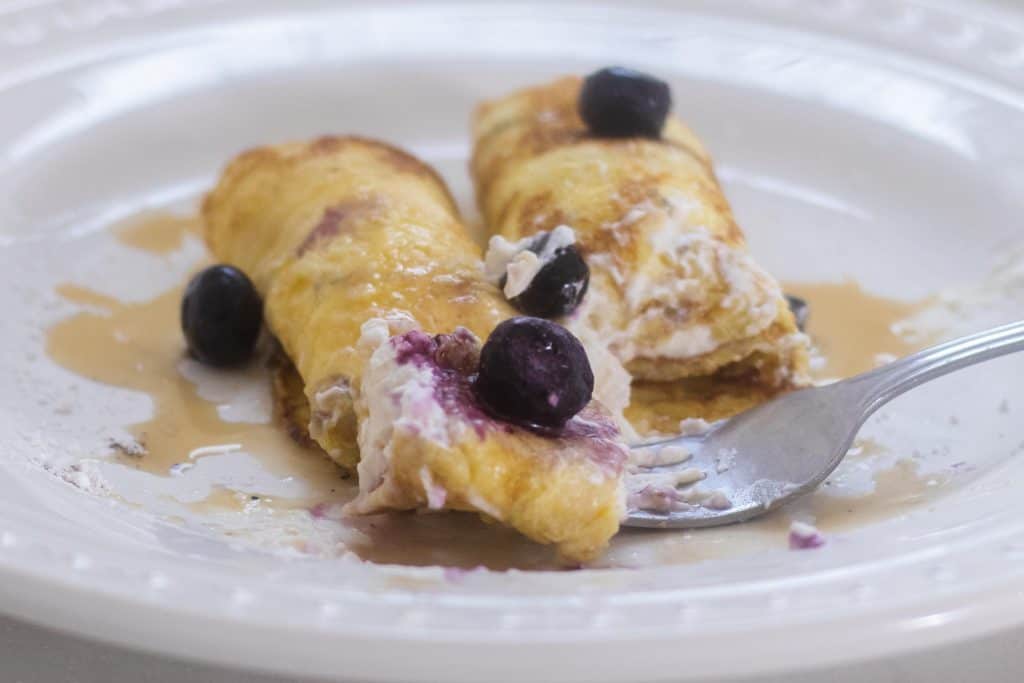 sourdough crepes with a whipped cream cheese filling rolled up and toped with blueberries on a white plate