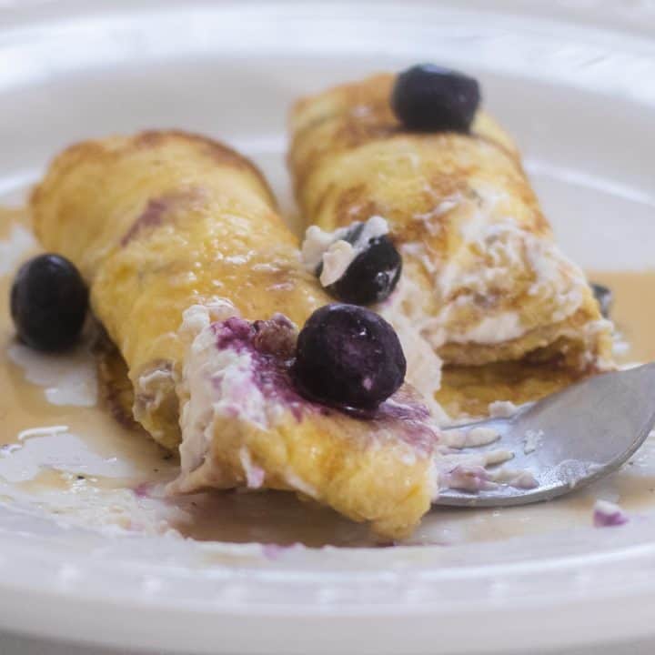 sourdough crepes with a whipped cream cheese filling rolled up and toped with blueberries on a white plate