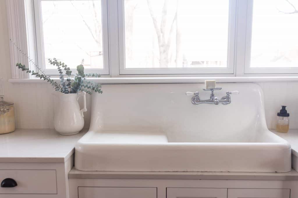 vintage white farmhouse apron sink with a beautiful chrome faucet. Fresh eucalyptus in a vintage iron stone vase to the right on quarts countertops