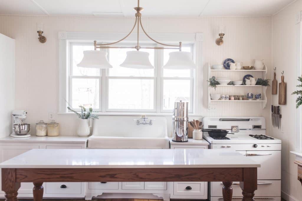 victorian farmhouse kitchen with long work table with quarts countertop. 3 pendant brass light fixture hangs over the table