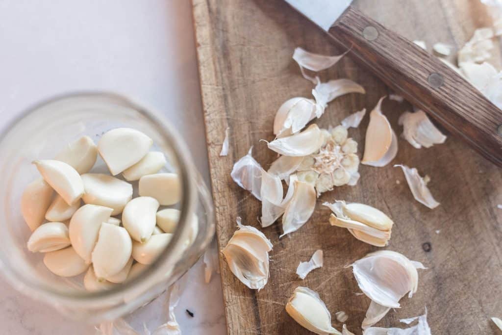 cutting board of garlic peels and a knife with a jar of whole peeled garlic cloves to the left.