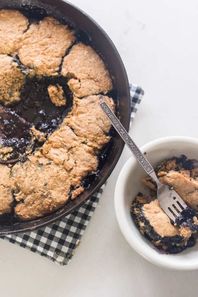 sourdough blueberry cobbler in a cast iron skillet. A scoop of cobbler to the right in a white dish.