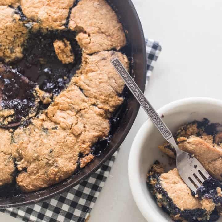 sourdough blueberry cobbler in a cast iron skillet. A scoop of cobbler to the right in a white dish.