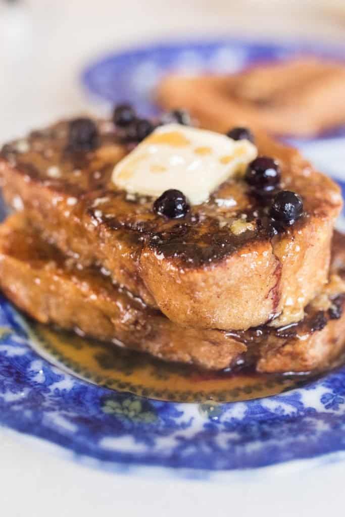 a vintage blue plate with two pieces of sourdough French toast stacked up and topped with butter, maple syrup, and blueberries. Another place of French toast in the background