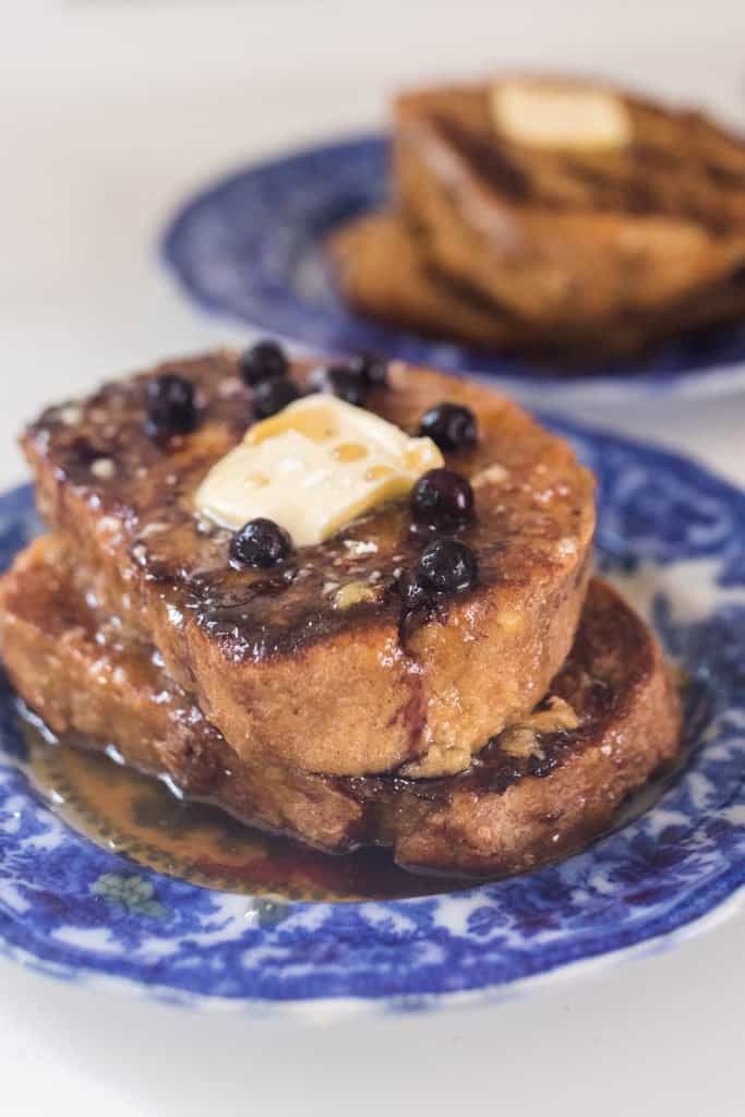 two plates with two pieces of sourdough French toast topped with blueberries, butter and maple syrup