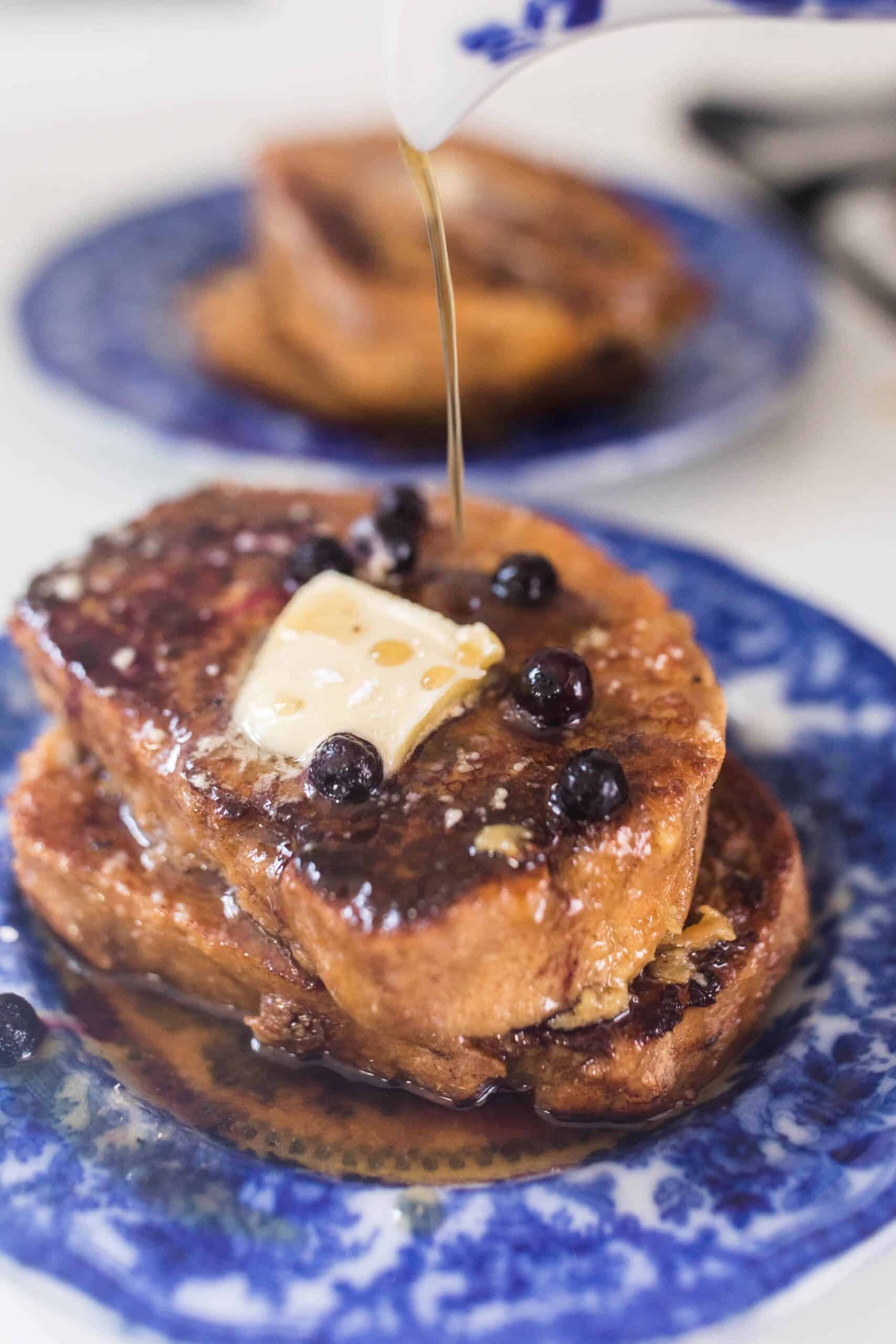 maple syrup being poured over sourdough French toast with blueberries on a blue plate with another plate of French toast in the background