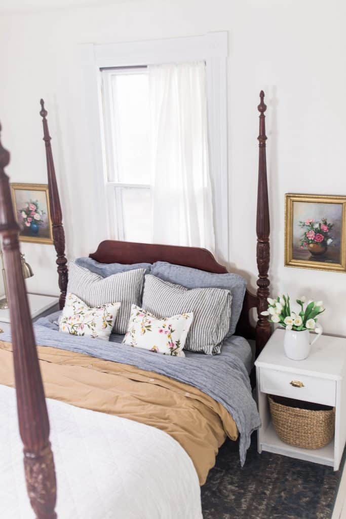 Victorian four poster bed with white, gold and blue stripped linens. Linen pillows are stacked against the headboard. To the right of the bed is a white nightstand with vase of flowers on top and gold framed art