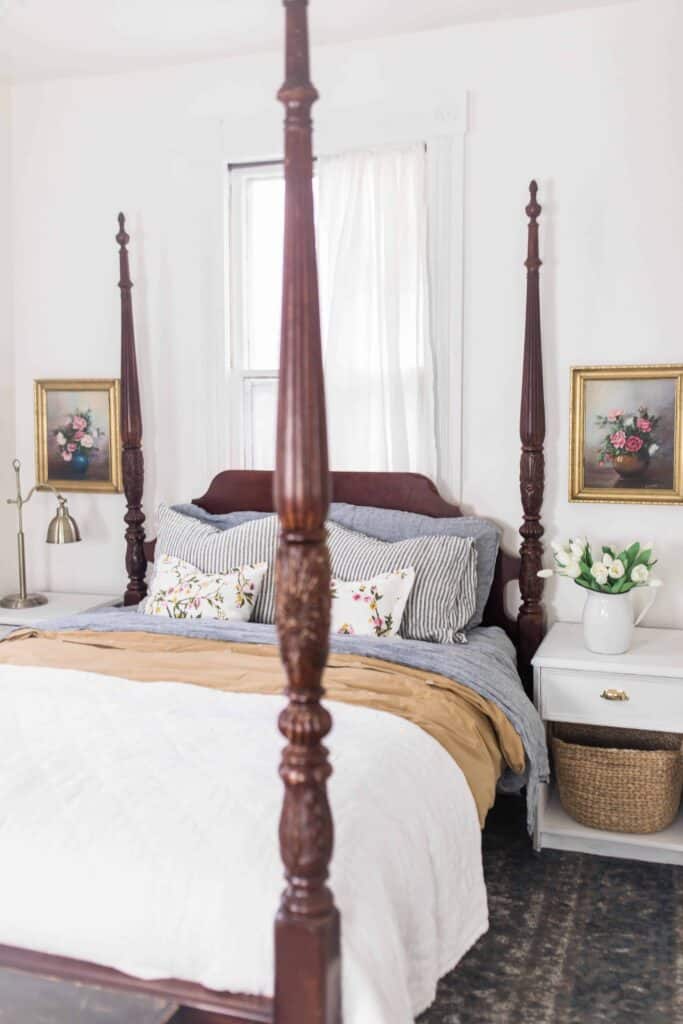 Beautiful dark wood four poster bed draped with linen bedding. White nightstands with a bottom basket to the right. Antique floral pictures with gold frames hang over the nightstands