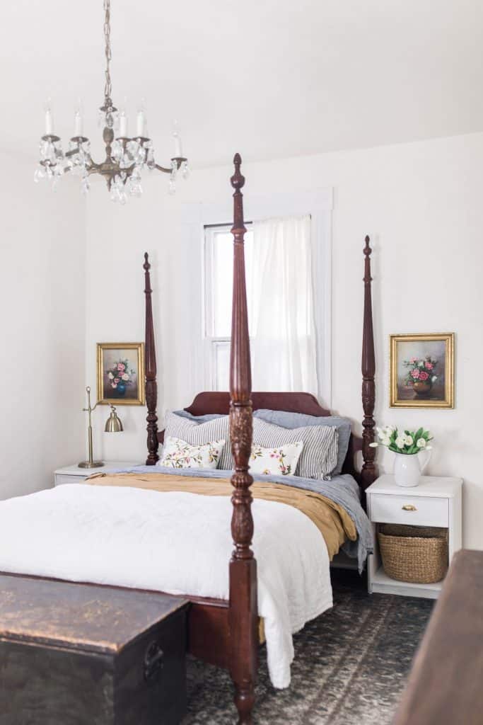 victorian farmhouse master bedroom with a four poster bed with white, gold, and blue linens, a chest at the foot of the bed and white nightstands to each side. Antique pictures hand above the nightstands