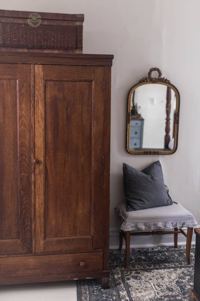 Large wood armoire with a woven chest on top. A footstool with ruffle slipcover and pillow to the right. A mirror hanging over the bench