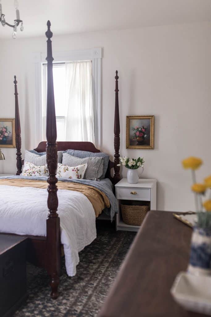 side view of a four poster bed with white, gold, and blue linens. Linen striped and floral pillows stacked on the bed. The top of a dresser with yellow florals in a vase to the side