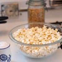 homemade coconut oil popcorn in a glass bowl on a stove top. popcorn kernels in the behind the bowl