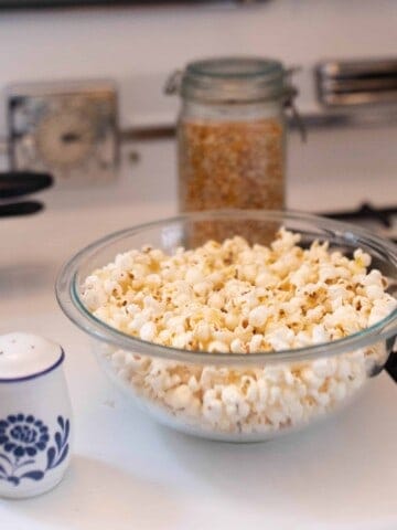 homemade coconut oil popcorn in a glass bowl on a stove top. popcorn kernels in the behind the bowl