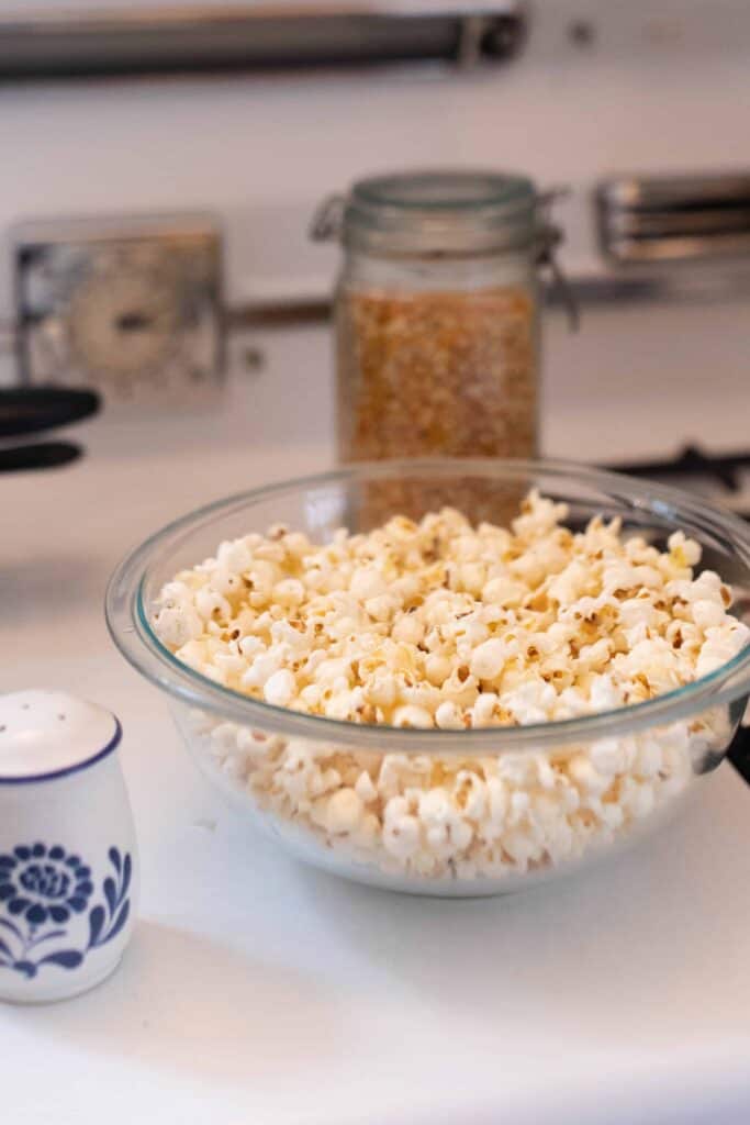 homemade coconut oil popcorn in a glass bowl on a stove top. popcorn kernels in the behind the bowl