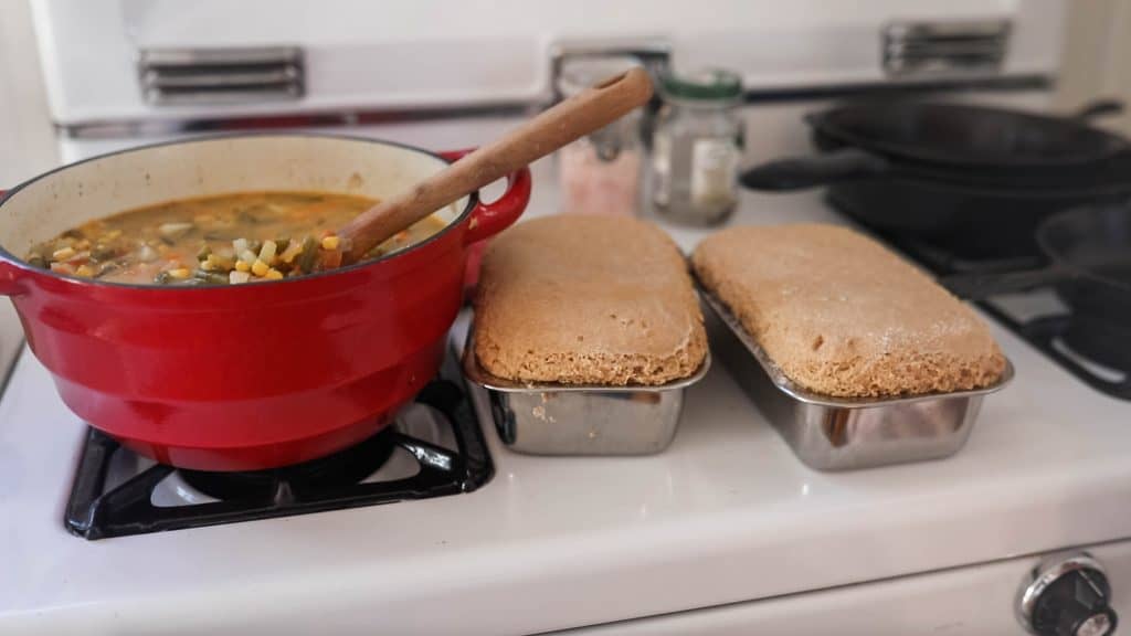 two loaves of einkorn bread in loaf pans next to a pot of soup on the oven