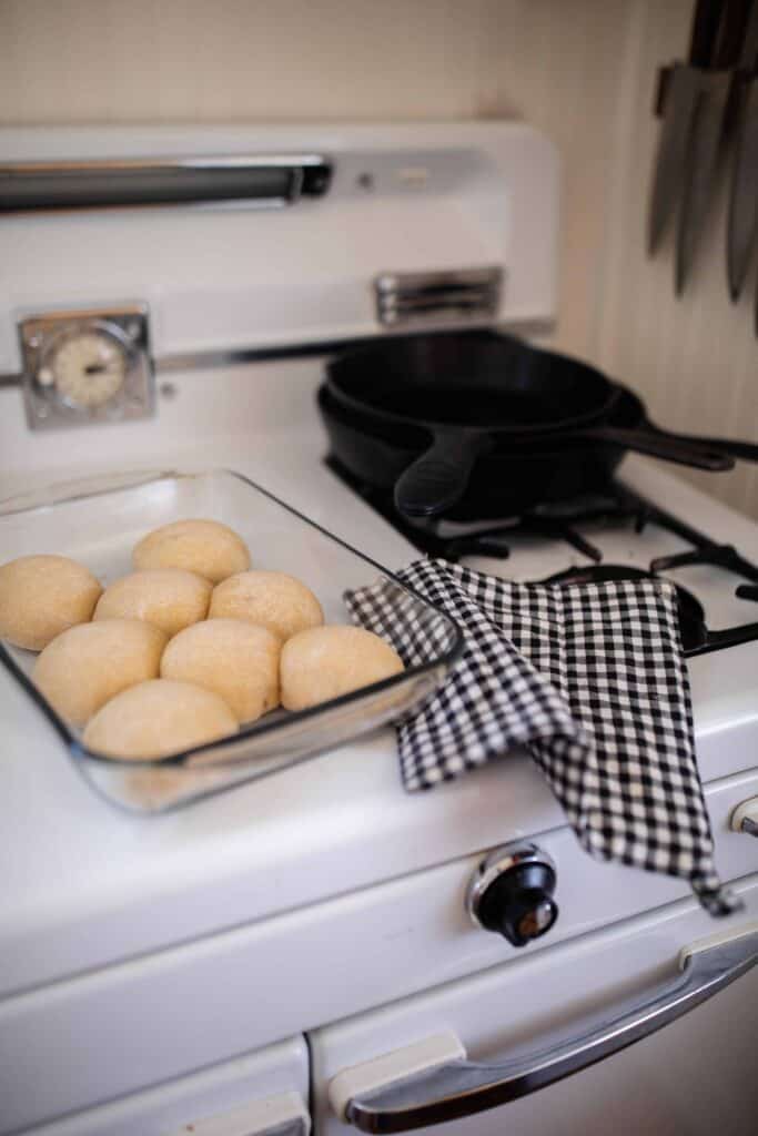 sourdough rolls in a baking dish on top a antique oven with a blue and white plaid towel to the right and cast iron skillet in the background.