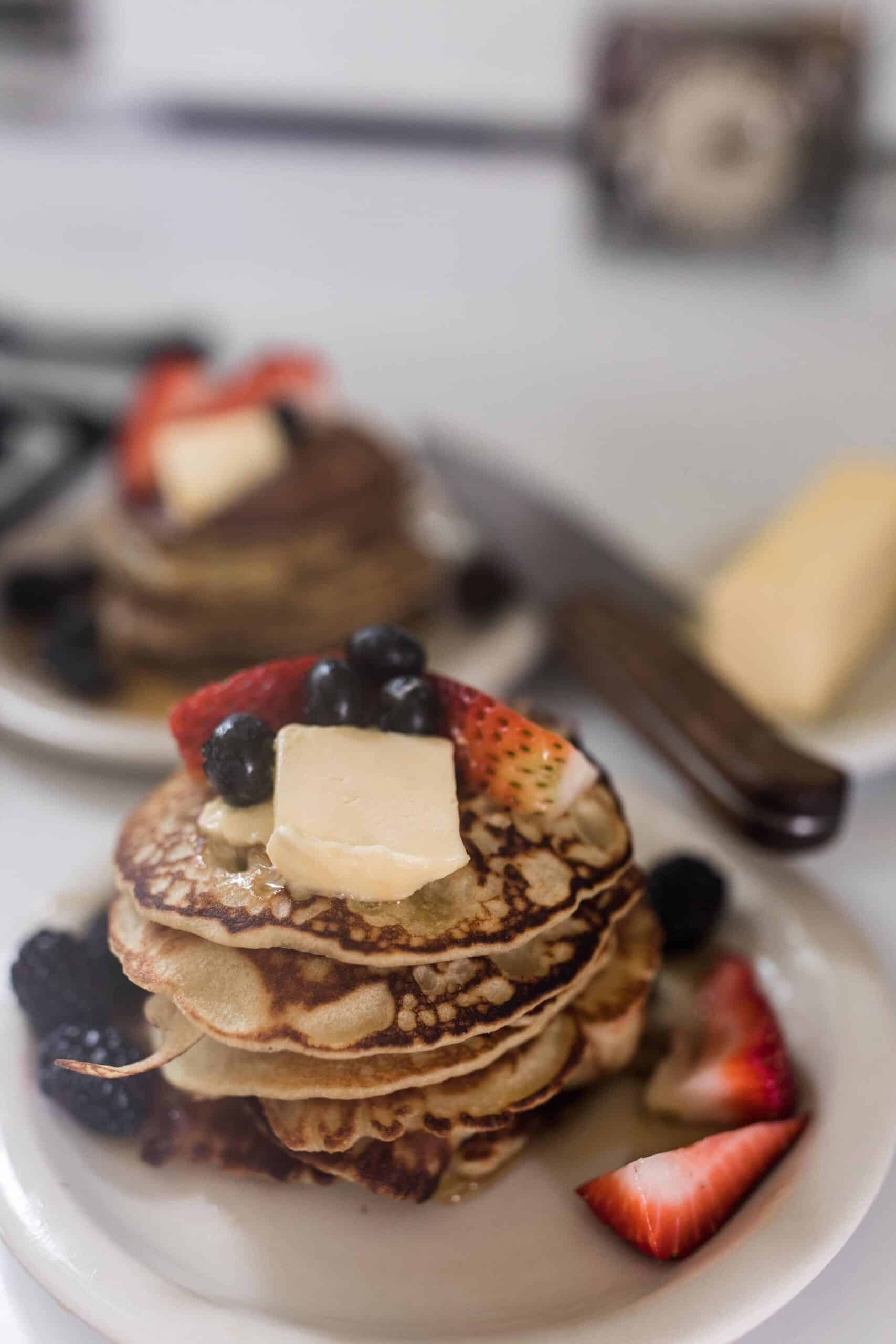 two stacks of buckwheat pancakes topped with fresh fruit and butter on white places