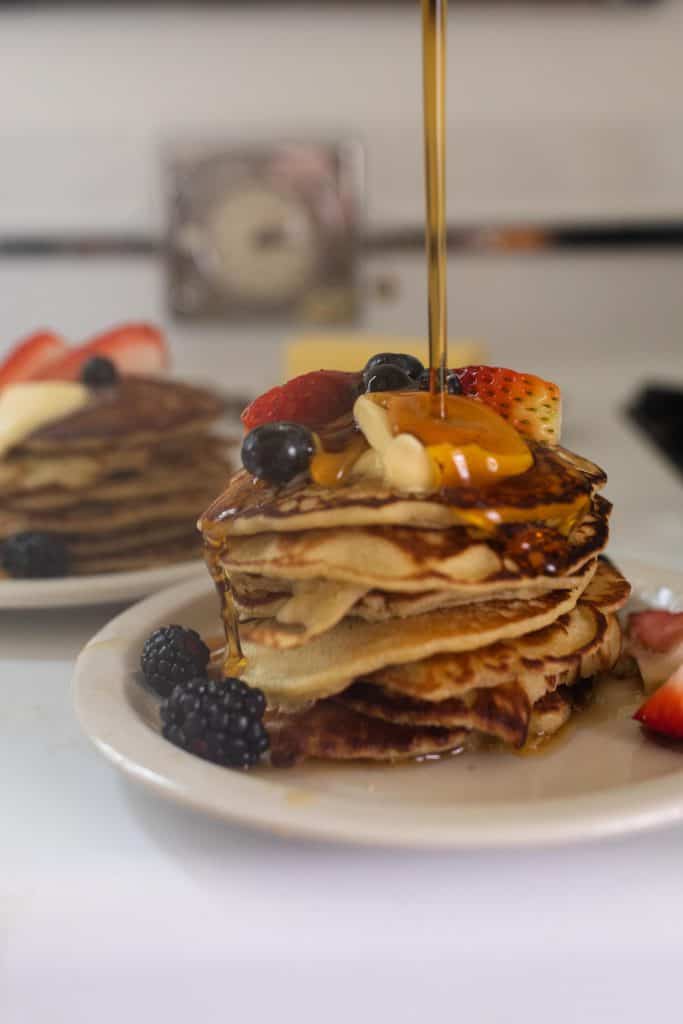 maple syrup being poured over a stack of buckwheat pancakes topped with butter and fruit