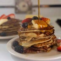 maple syrup being poured over a stack of buckwheat pancakes topped with butter and fruit