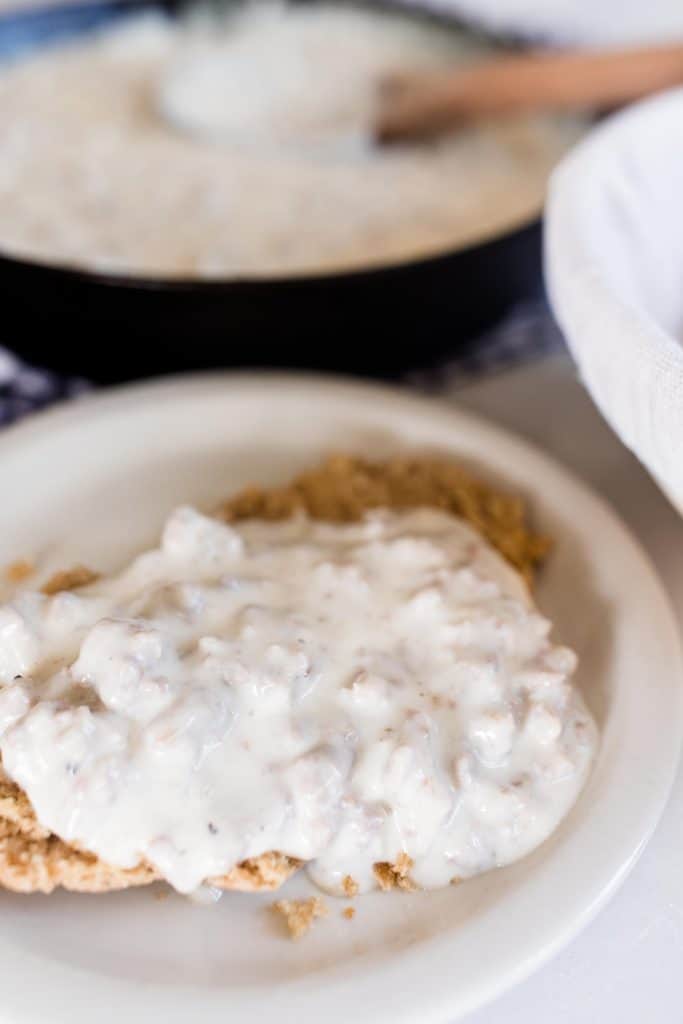 sourdough biscuits and gravy on a white plate with a cast iron skillet of sausage gravy in the background