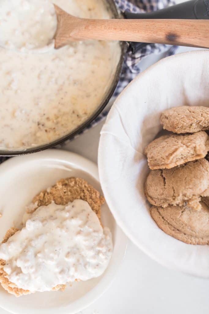 sourdough biscuits and gravy on a white plate. A white bowl of biscuits are to the right and a cast iron skillet with sausage gravy in the background