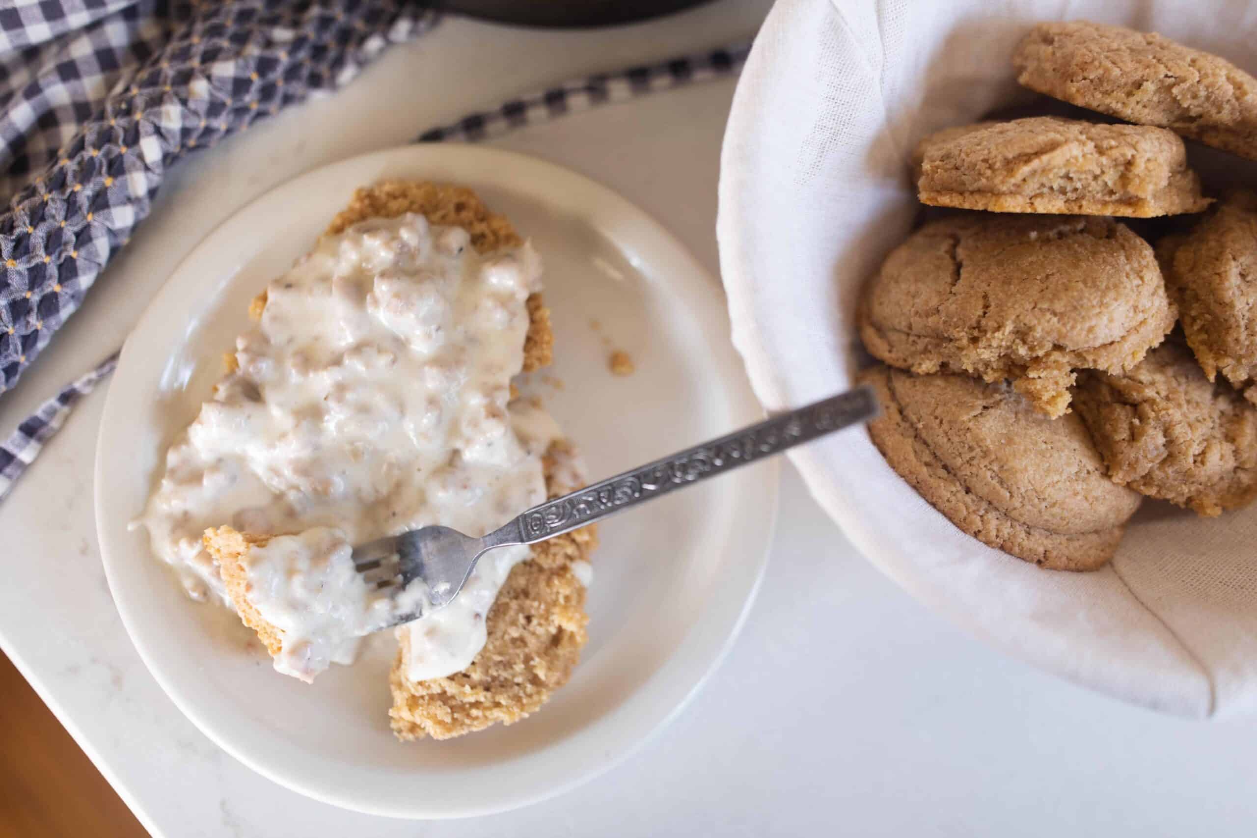 sourdough biscuits smothered in sausage gravy on a white plate. A bowl of sourdough biscuits to the right