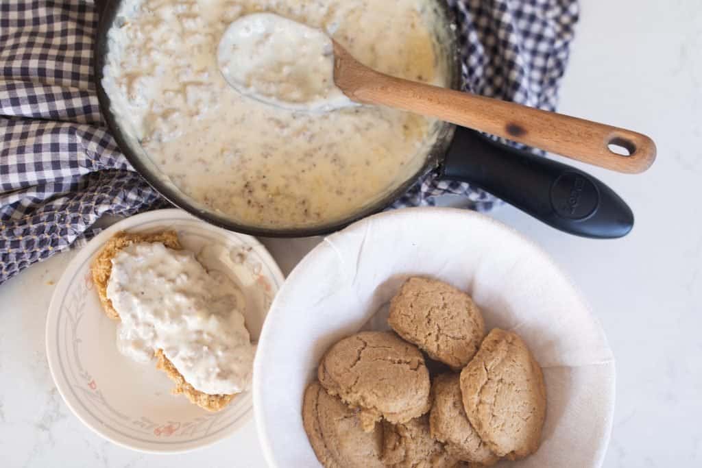 bowl of sourdough biscuits. A cast iron skillet with sausage gravy sits on a white and blue checked towel and a plate of biscuits covered in gravy is next to the bowl
