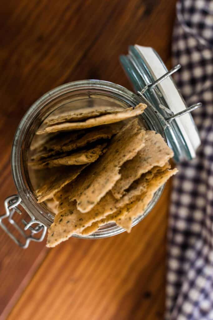 overhead shot of homemade sourdough crackers in a glass jar with lid. A white and blue checked towel is to the right