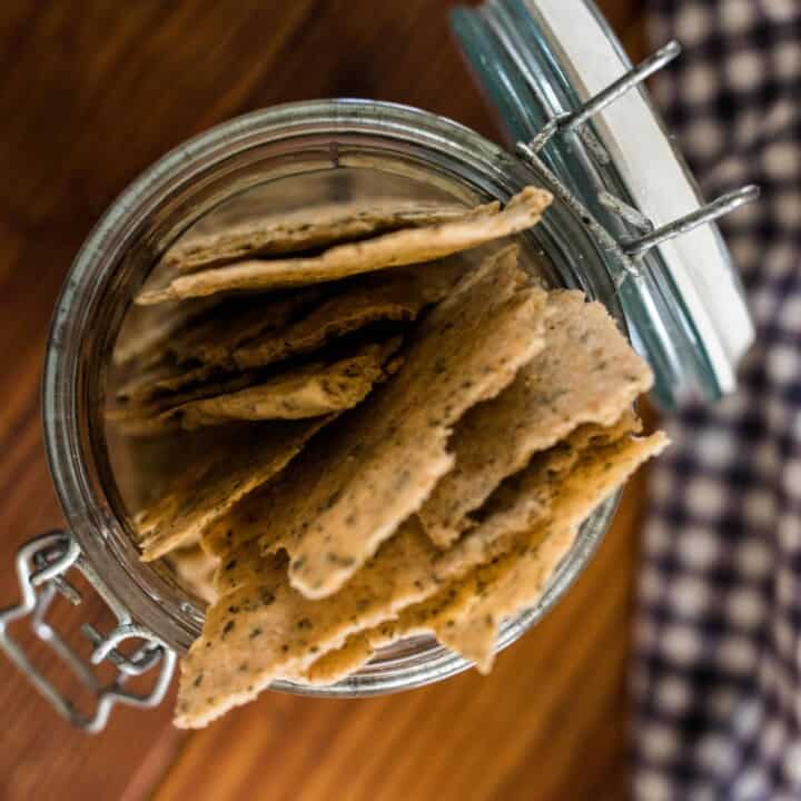 overhead shot of homemade sourdough crackers in a glass jar with lid. A white and blue checked towel is to the right