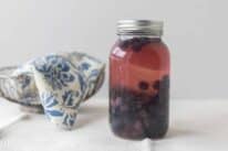 Fermented blueberries in a mason jar with a silver bowl covered with a blue and white damask printed towel in the background