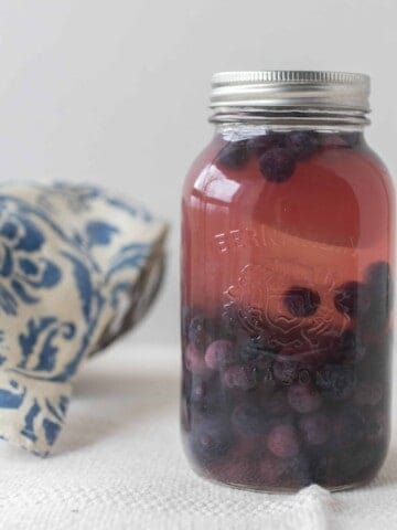 Fermented blueberries in a mason jar with a silver bowl covered with a blue and white damask printed towel in the background