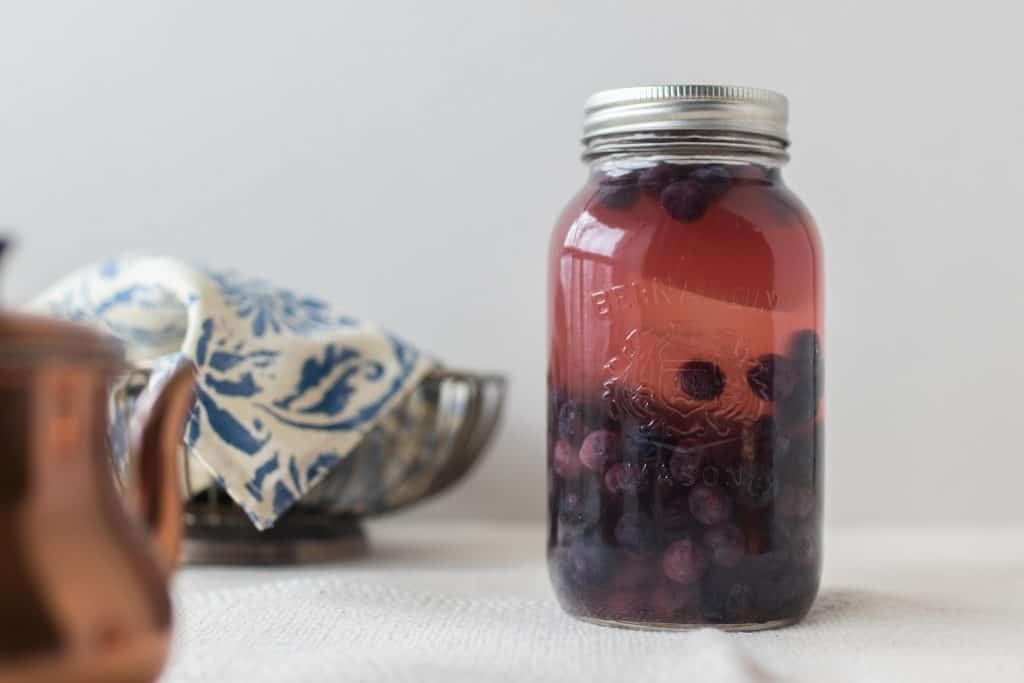 fermented blueberries in a mason jar with a copper tea pot and bowl covered with a blue and white napkin to the left