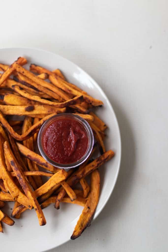 fermented ketchup in a glass jar surrounded by sweet potato fries on a white plate