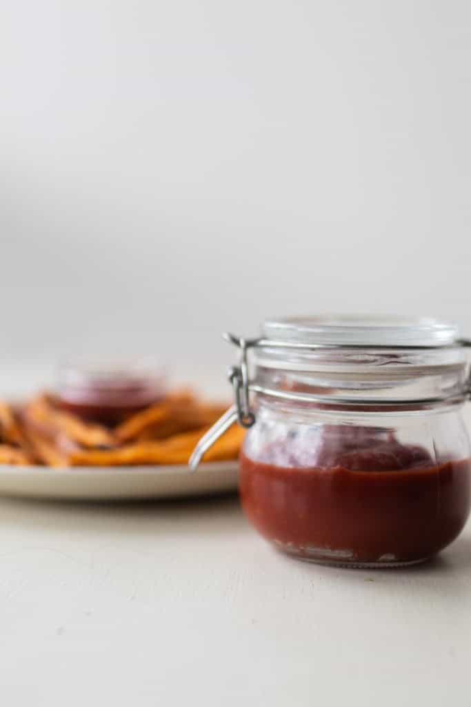 a swing top jar full of homemade ketchup with a plate of sweet potato fries in the background