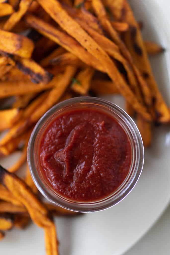 homemade ketchup in a jar on a white plate with sweet potato fries