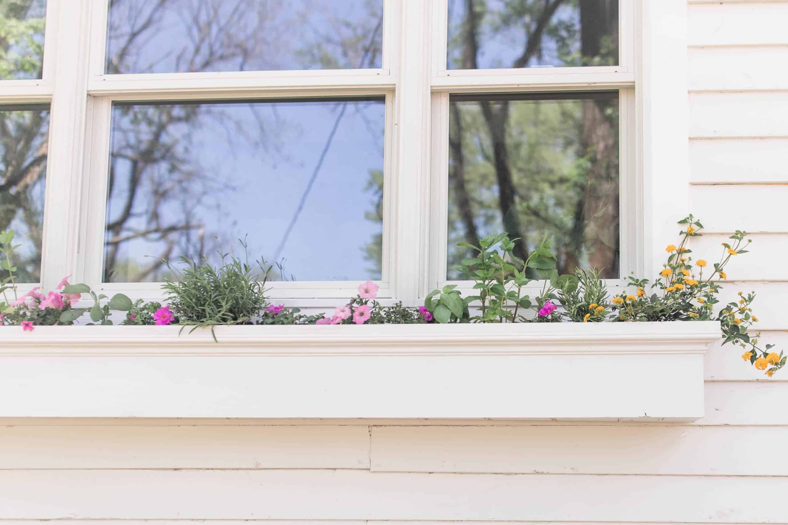 a white window box hanging below a window on a white farmhouse with pink and yellow flowers in it.