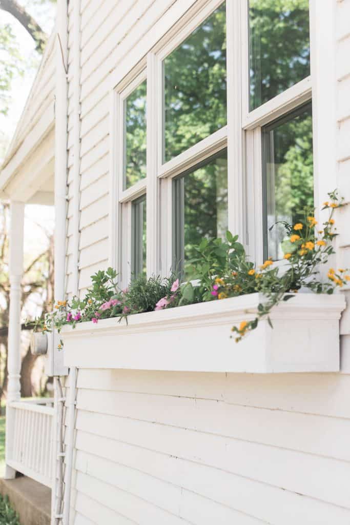 a diy window planter box with yellow and pink flowers hanging below a window on a white farmhouse