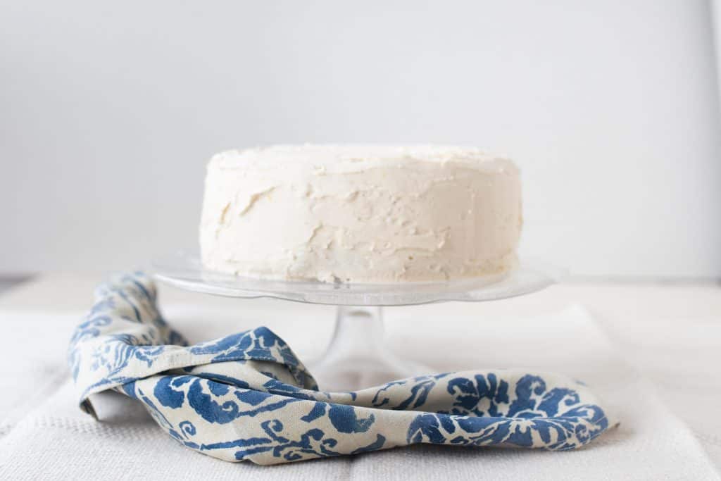 two tiered cake with white icing on a glass cake stand. A blue and white damask napkin is laying underneath the cake stand