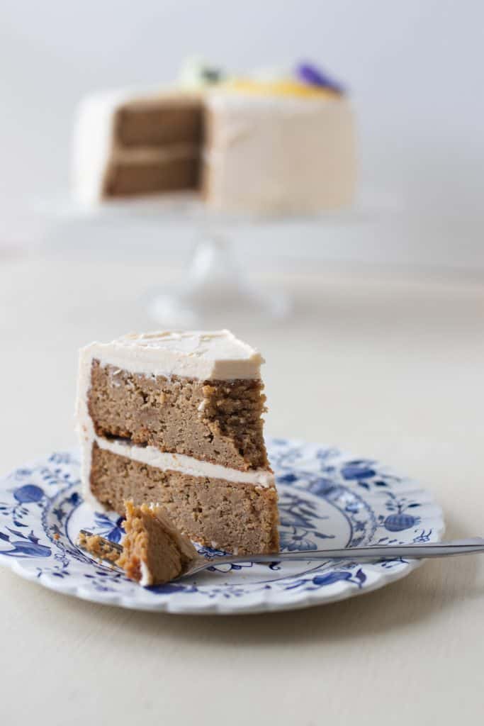 a slice of two tiered gluten free lemon cake with earl grey and lemon icing on a blue and white plate with a fork. The remaining cake sits on a cake stand in the background