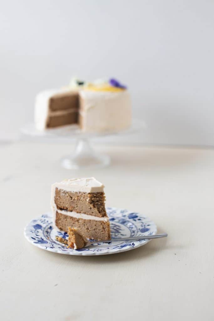 slice of two tiered lemon cake with white icing on an antique blue and white plate with a fork. The remaining cake on a cake stand and topped with lemon slices and purple edible flowers is in the background.