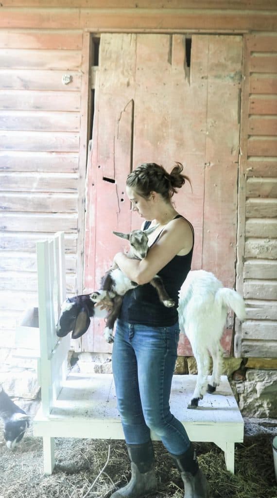 women holding baby goat with a momma goat in the background standing on a goat stand.
