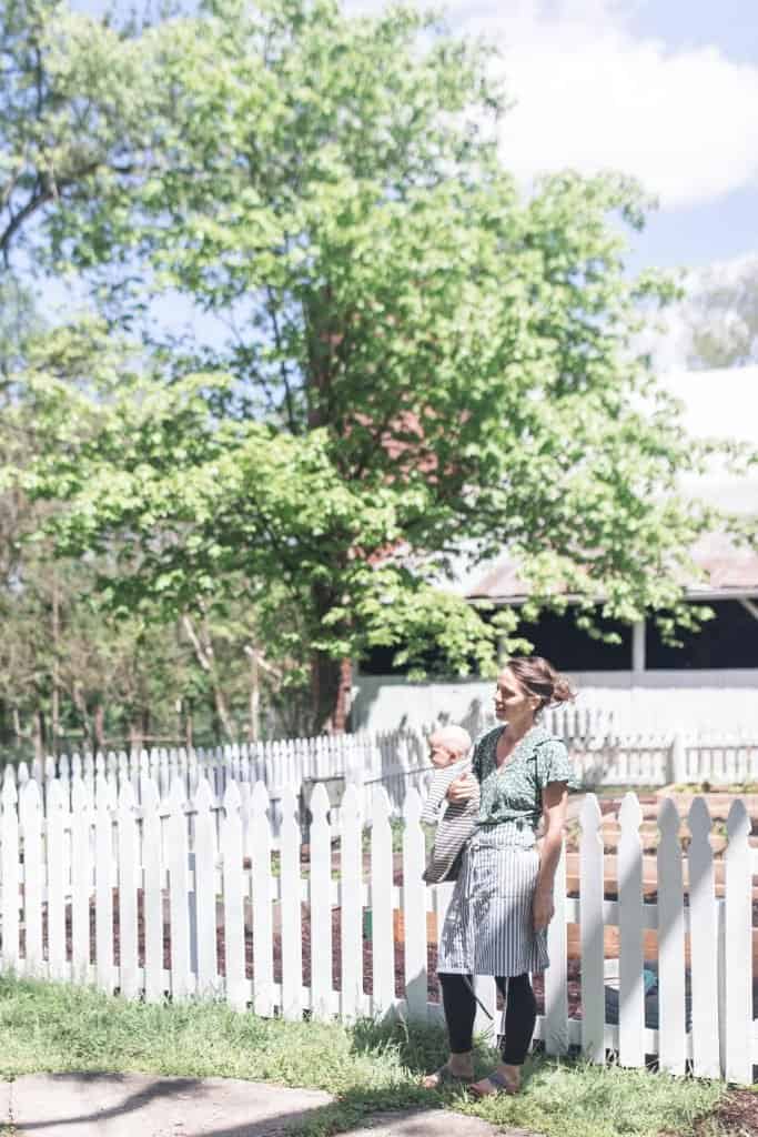 women wearing a green dress and an apron holding a baby on her hip standing in front of a DIY picket fence