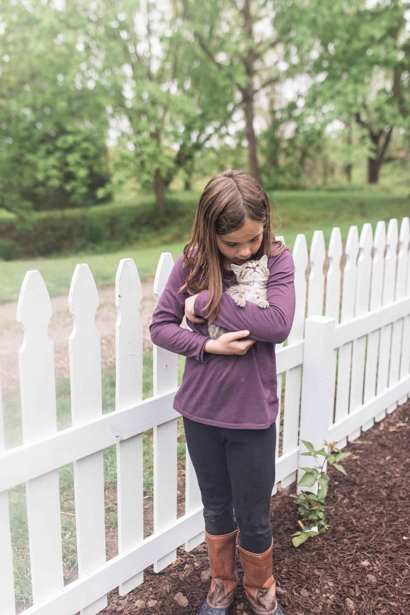 girl wearing a purple shirt cuddling a kitten in front of a DIY picket fence