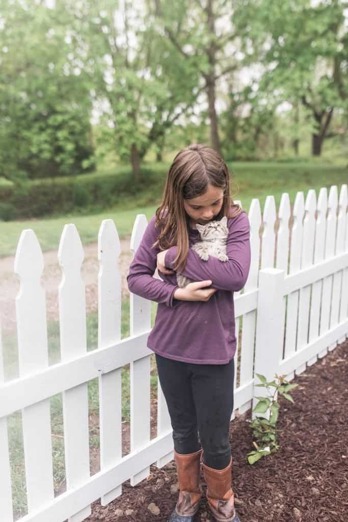 girl wearing a purple shirt cuddling a kitten in front of a DIY picket fence