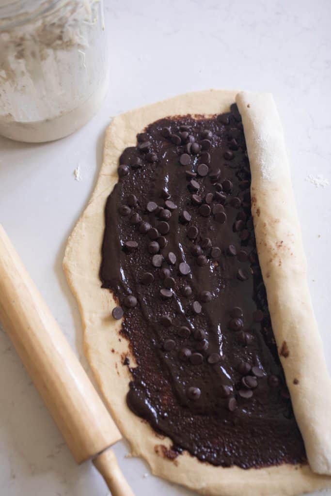 chocolate and sugar mixture spread out on sourdough dough and sprinkled with chocolate chips. The dough is starting to be rolled up halfway